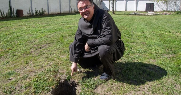 A smiling monk in a black robe kneels near a hole in a grassy field, wearing glasses and sandals.
