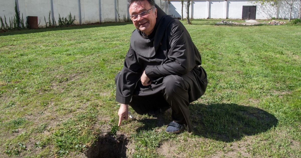 A smiling monk in a black robe kneels near a hole in a grassy field, wearing glasses and sandals.