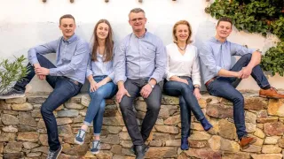 A family of five sitting on a stone wall, smiling. Two men and three women, all wearing light blue shirts. The wall is white, and there is a plant in the background.