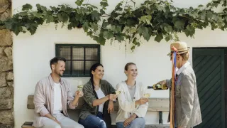Three individuals seated on a bench, one holding a wine bottle, while two others have wine glasses, smiling and posing for a photo.