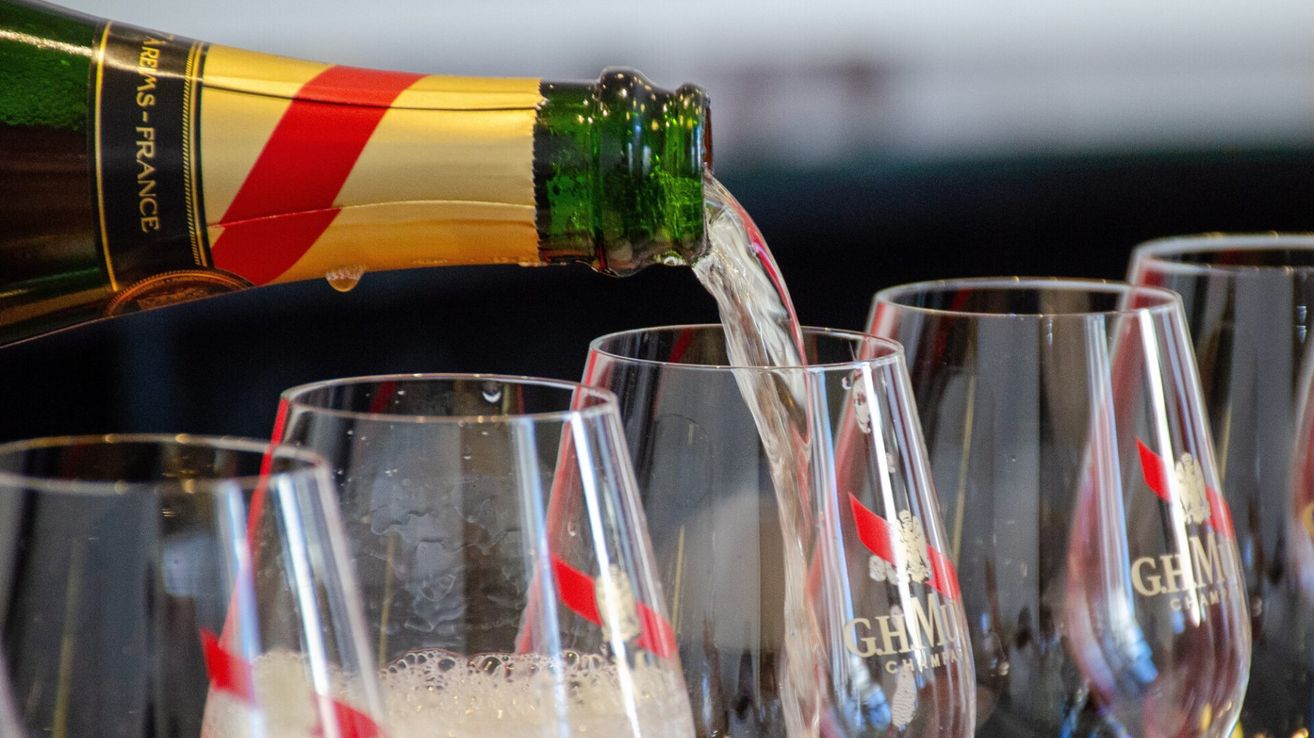 Champagne is being poured into glasses with red and white ribbons. The bottle has a green cap and the wine glasses have the GHMU brand logo.