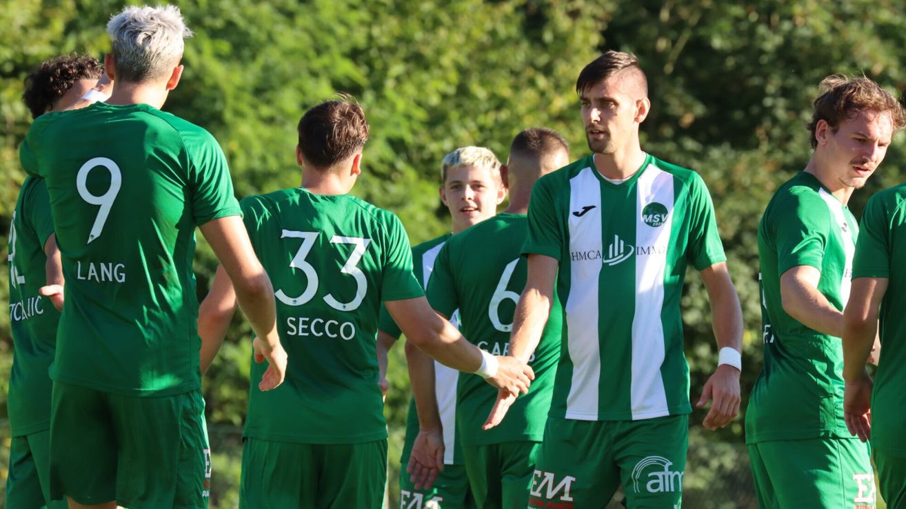 A group of soccer players in green uniforms converse outside. Player 33 from SECCO is in the foreground, while another player with the number 6 stands behind him.
