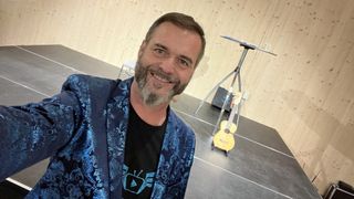 A man with a beard smiles in front of a wooden wall. He wears a blue jacket and black t-shirt. Behind him is a small guitar on a stand.