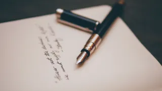 A fountain pen is placed on a piece of paper with handwritten text. The pen has a black body and a gold tip. The paper has a white background and cursive handwriting.
