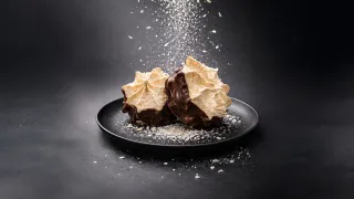 Two chocolate-coated meringues with almond flakes on a black plate, dusted with powdered sugar, captured in a moment of sweetness.