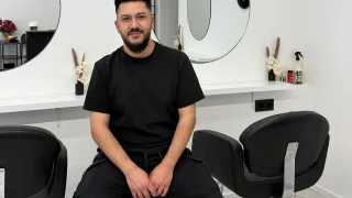 A man sits in a chair at a hair salon. He wears a black shirt and pants. Mirrors and a white shelf are behind him.