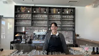 A woman in a black apron stands behind a bar counter, surrounded by glasses, bottles, and an espresso machine, in a room with a brick wall.