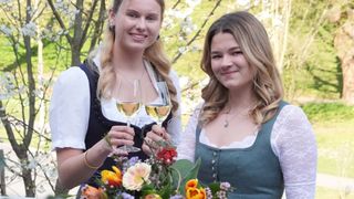 Two young women, dressed in traditional attire, are smiling and holding wine glasses. They are surrounded by a bouquet of flowers, and the background features a garden with trees and greenery.