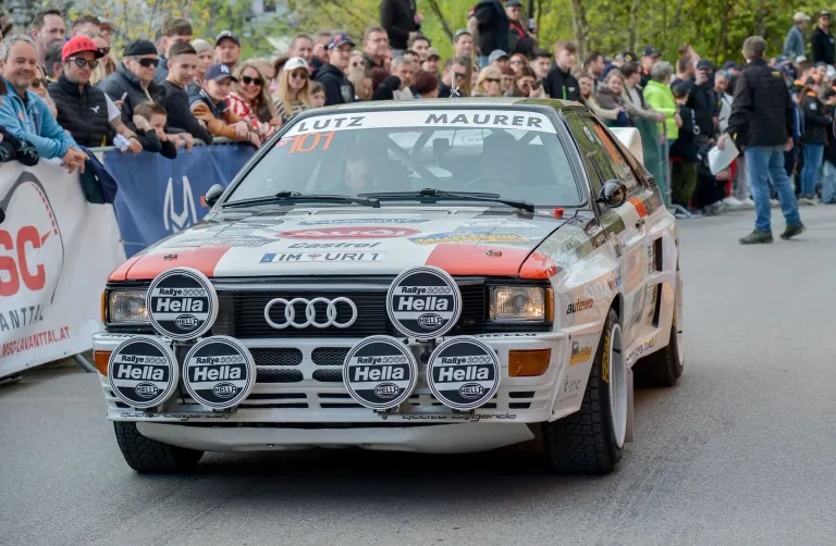 A rally car with the number 101 is driving on the street, surrounded by a crowd of people. The car has multiple Hella badges on its front bumper.