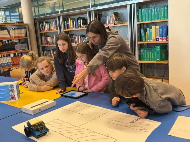 A group of children, guided by a woman in glasses, are gathered around a blue table, engaged in a project involving a tablet, a toy robot, and a drawing.