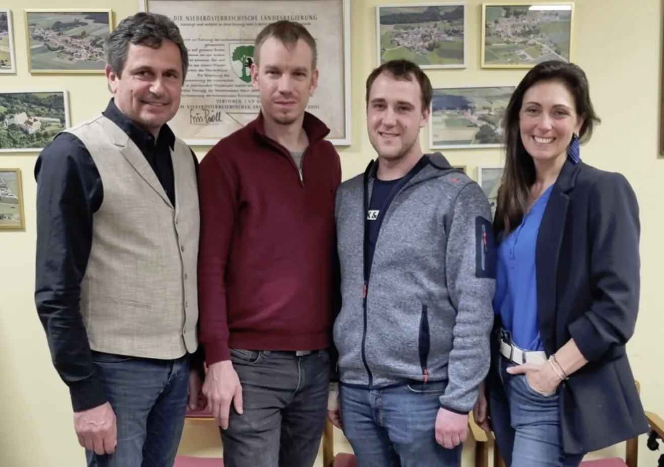 Four people are smiling in a room with a yellow wall. Two men are wearing jackets and jeans, one woman wears a blue shirt, and another man wears a beige jacket.