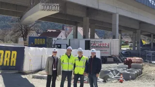 Four men in high visibility vests stand under a bridge under construction. A truck and building are in the background.