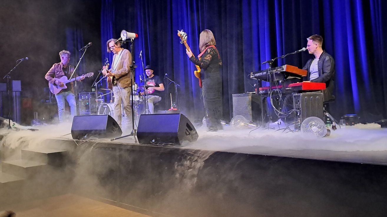 A band is performing on stage with fog and blue curtains. A man plays guitar, a woman plays guitar and sings, and a man plays drums.