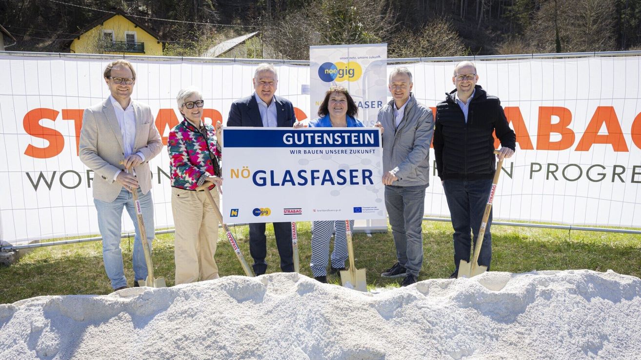 A group of people holding shovels, standing behind a sign, which reads Gutenstein No Glasfaser. The man on the right is wearing glasses and a jacket.