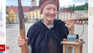 An elderly woman in medieval attire holds a staff and lantern, smiling in a town square with historic buildings in the background.