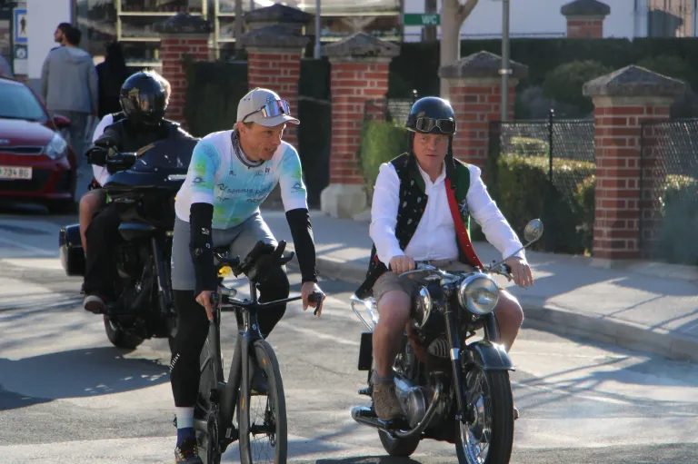 Three men on bikes, one riding a bicycle, another on a motorcycle, and a third on a motorcycle in the background, all wearing helmets.