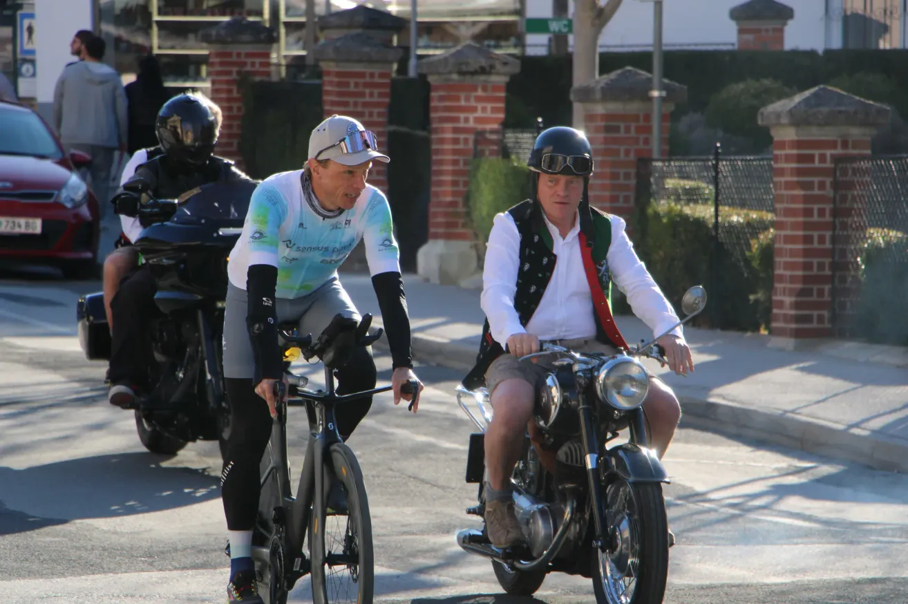 Three men on bikes, one riding a bicycle, another on a motorcycle, and a third on a motorcycle in the background, all wearing helmets.