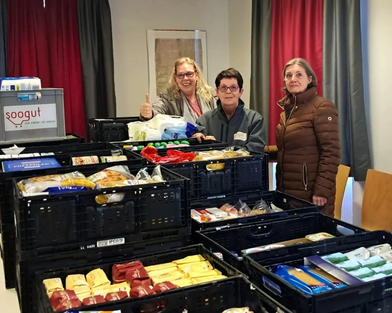 Three women stand next to crates filled with food. The woman in the middle gives a thumbs up. The woman on the right wears a brown jacket.