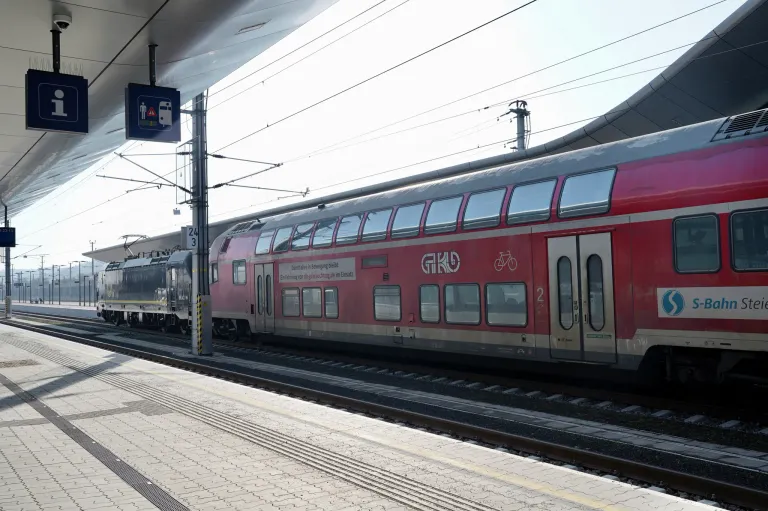 A red train with several windows is parked on the tracks at a station. A blue signboard is on the left side. The platform has a brick floor.