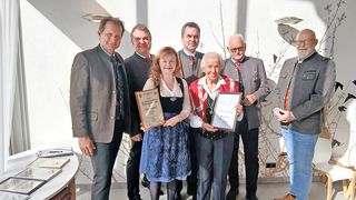 Seven people are standing and smiling for a photo. Two women are holding framed certificates, and the man on the right is wearing a white beard. The wall behind them has tree-like decorations. A desk is on the left with papers and a pen.