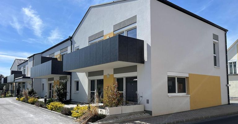 Ein modernes zweistöckiges Haus mit Balkon, Glasfenstern und grau-gelbem Äußeren. Es verfügt über einen gepflasterten Weg, Pflanzen und einen klaren blauen Himmel.