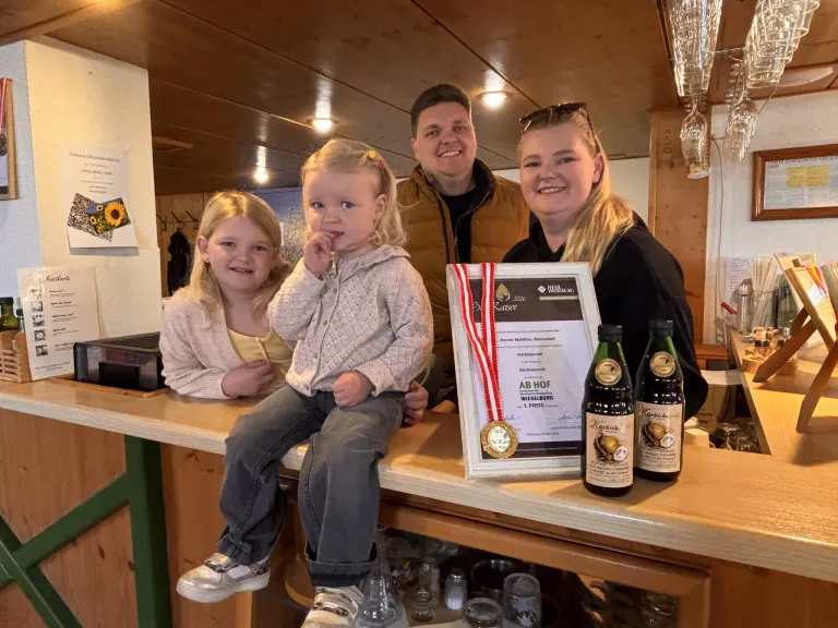 Eine vierköpfige Familie posiert mit einem jungen Mädchen, das eine Medaille trägt, an einer Bar. Sie lächeln und halten ein Zertifikat, mit einer Weinflasche und einem Glas in der Nähe.
