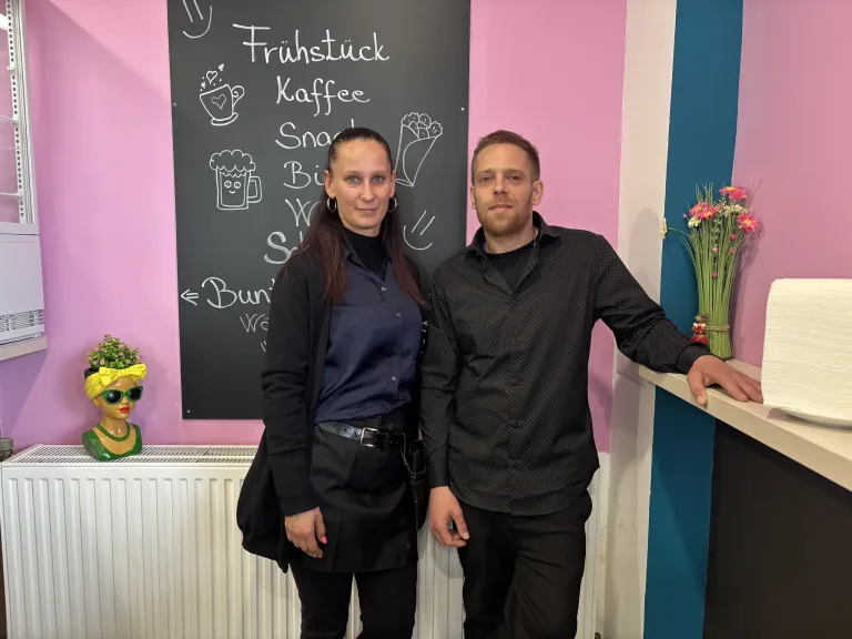 A man and a woman stand smiling in front of a blackboard with drawings. The woman is wearing a black coat, and the man has his hand on the counter.
