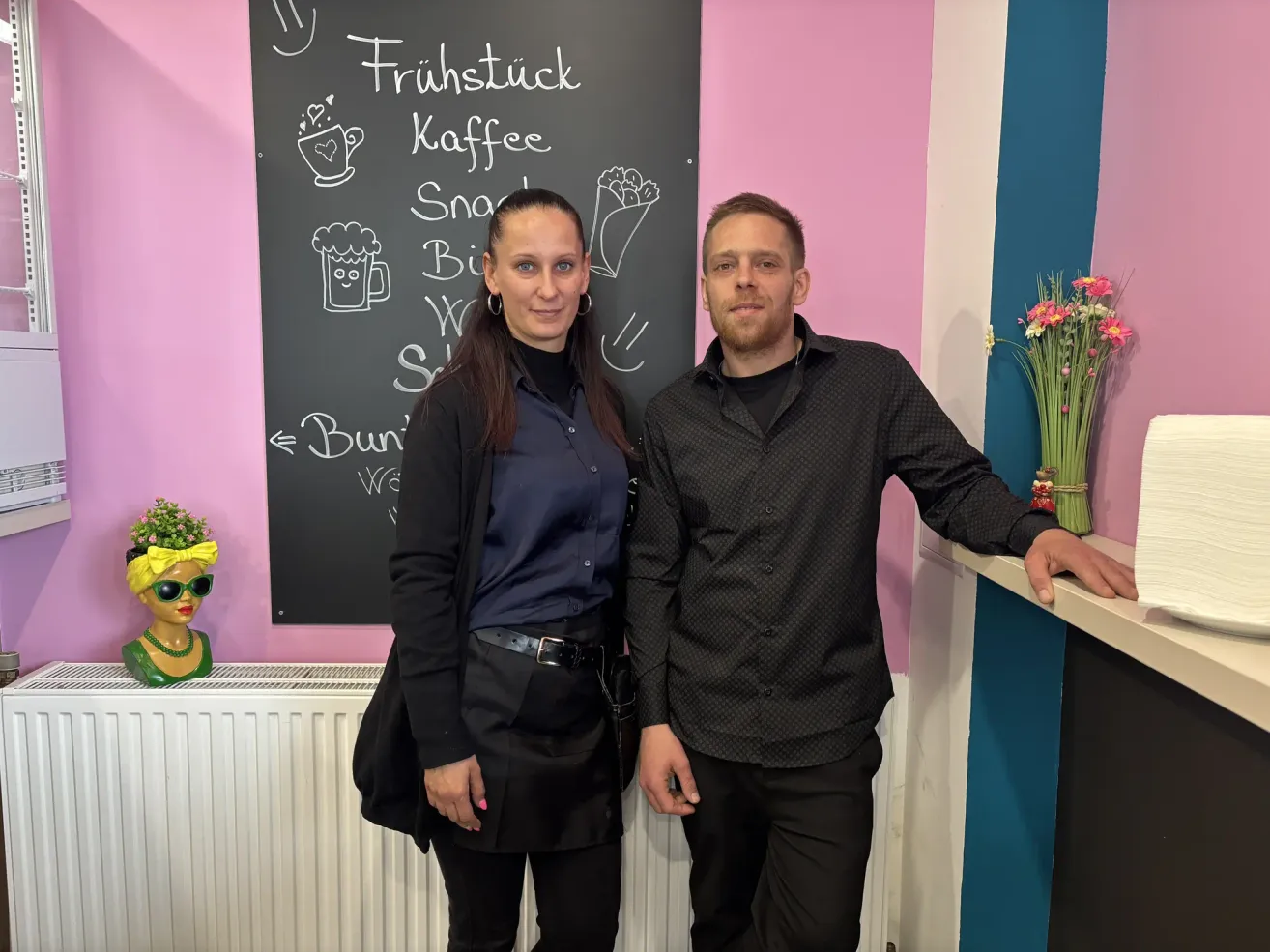 A man and a woman stand smiling in front of a blackboard with drawings. The woman is wearing a black coat, and the man has his hand on the counter.
