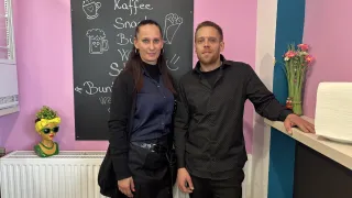 A man and a woman stand smiling in front of a blackboard with drawings. The woman is wearing a black coat, and the man has his hand on the counter.