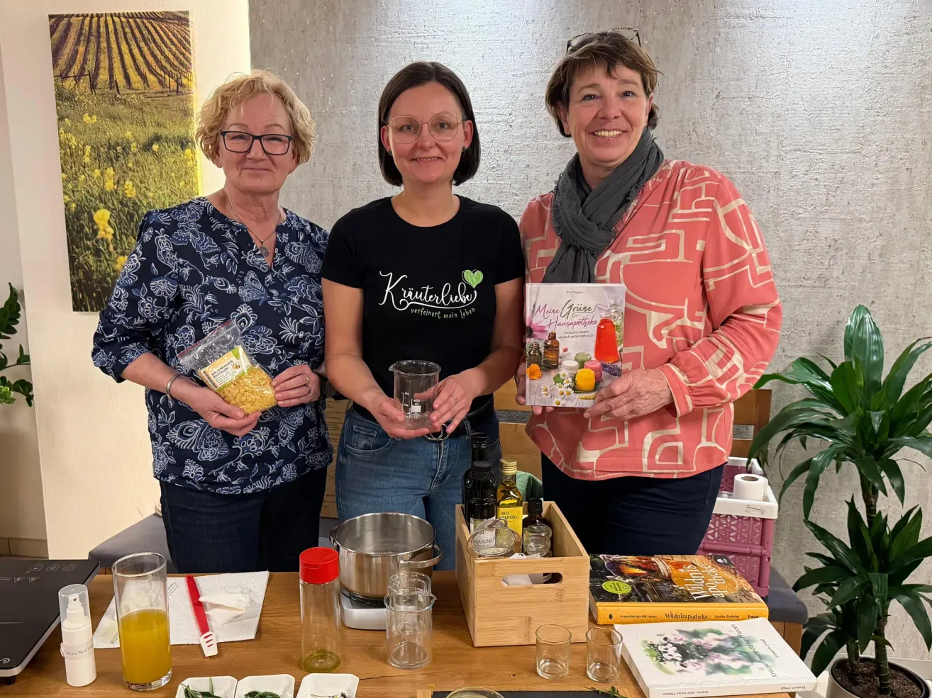 Three women are standing behind a table with a cooking pot, oil, and books. The woman on the left is holding a plastic bag of yellow beans. The woman in the middle is holding a cup. The woman on the right is holding a book.