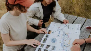 Four individuals sit around a picnic table outdoors, studying a map. Two women wear hats, and one wears glasses. The map has pictures and text.