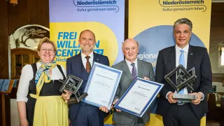 Four individuals hold awards in front of a backdrop with the text 'Frei Willen' and logos for 'Kultur. Region. Niederösterreich'. The man on the left wears glasses and a scarf. The others wear suits and ties. The man on the right wears a blue tie.