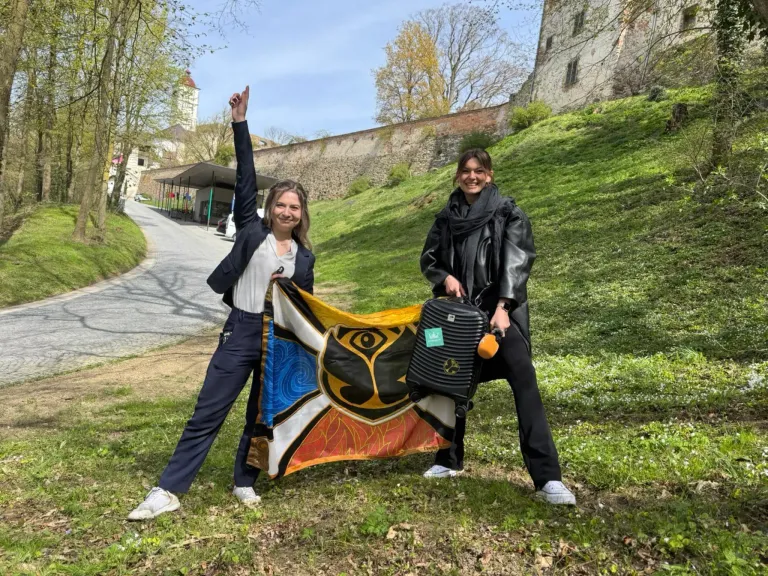 Two women stand on a grassy hill, one holding a colorful flag with a cat design. They smile and pose for a photo.