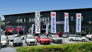 Multiple Suzuki cars parked outside a dealership, banners for Suzuki and KYCK in front, a clear sky above.