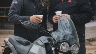Two men in hoodies standing next to a motorcycle, holding cups. The man on the left has a beard and the man on the right has a beanie.