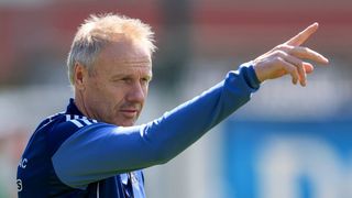 An older man with gray hair is wearing a blue long-sleeve shirt. He is pointing his right hand upward and seems to be giving instructions or directions.