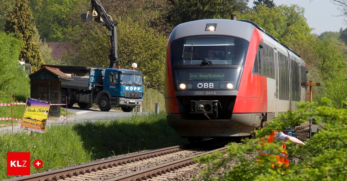 A blue truck with a crane drives past a red train on a sunny day. The train is heading to Graz Hauptbahnhof.