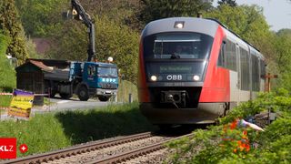 A blue truck with a crane drives past a red train on a sunny day. The train is heading to Graz Hauptbahnhof.