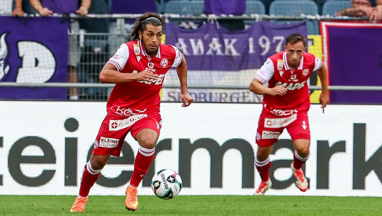 Two soccer players in red uniforms running on the field, one kicking a soccer ball. A banner in the background reads 'AWAK 1977'.