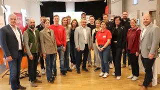 A group of people wearing formal and casual clothing stand together in a room with wooden floors, smiling and posing for a photo.