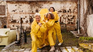 Three women in yellow rain gear pose for a photo in front of a stone wall with a metal post and plant nearby.