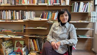 An older woman sits in front of a bookshelf filled with books, wearing a patterned shirt.