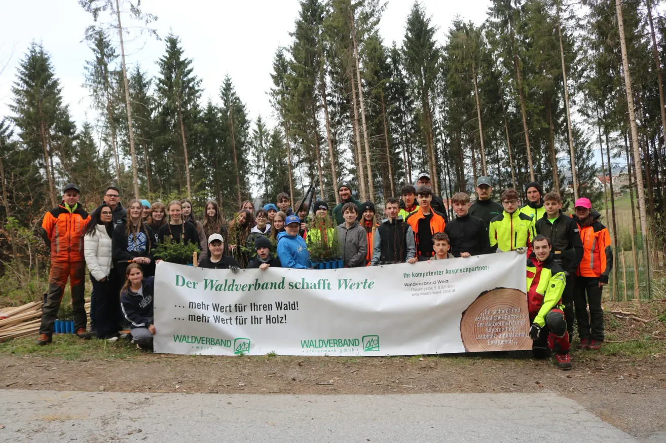 A group of people poses with a banner in a forest area. The banner reads 'Der Waldverband schafft Werte' and encourages value creation for forests and wood.