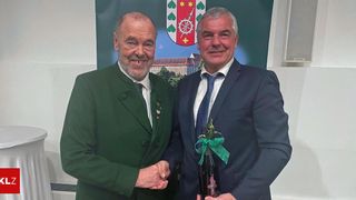Two men smile for a picture with one holding a wine bottle adorned with flowers and a ribbon. Behind them is a banner with a coat of arms.