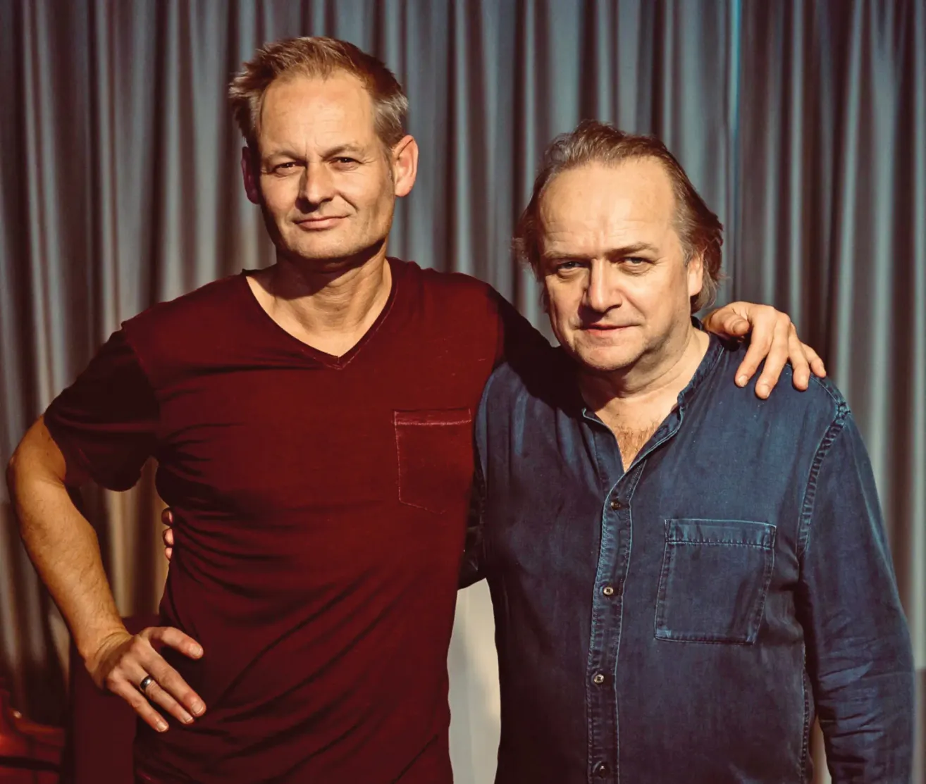 Two men, one wearing a red shirt and the other a blue shirt, standing close to each other with a smile. Behind them is a curtain.