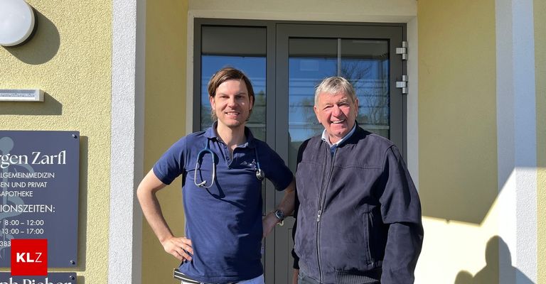 Two men stand in front of a door. The man on the left wears a stethoscope and a blue shirt. The man on the right wears a jacket and smiles.