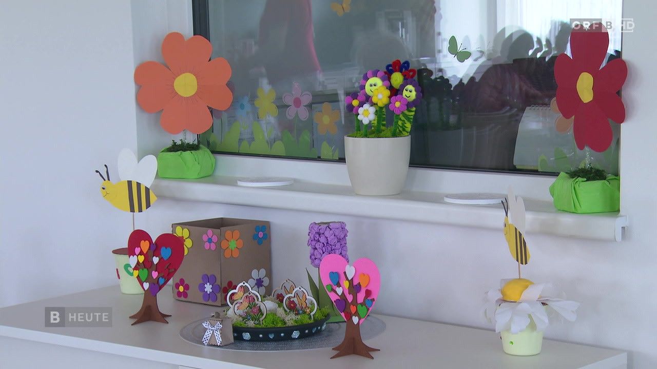 A windowsill displays colorful paper flowers and crafts. A heart tree with many hearts decorates the center. A flower vase and a heart-shaped box add to the cheerful decor.