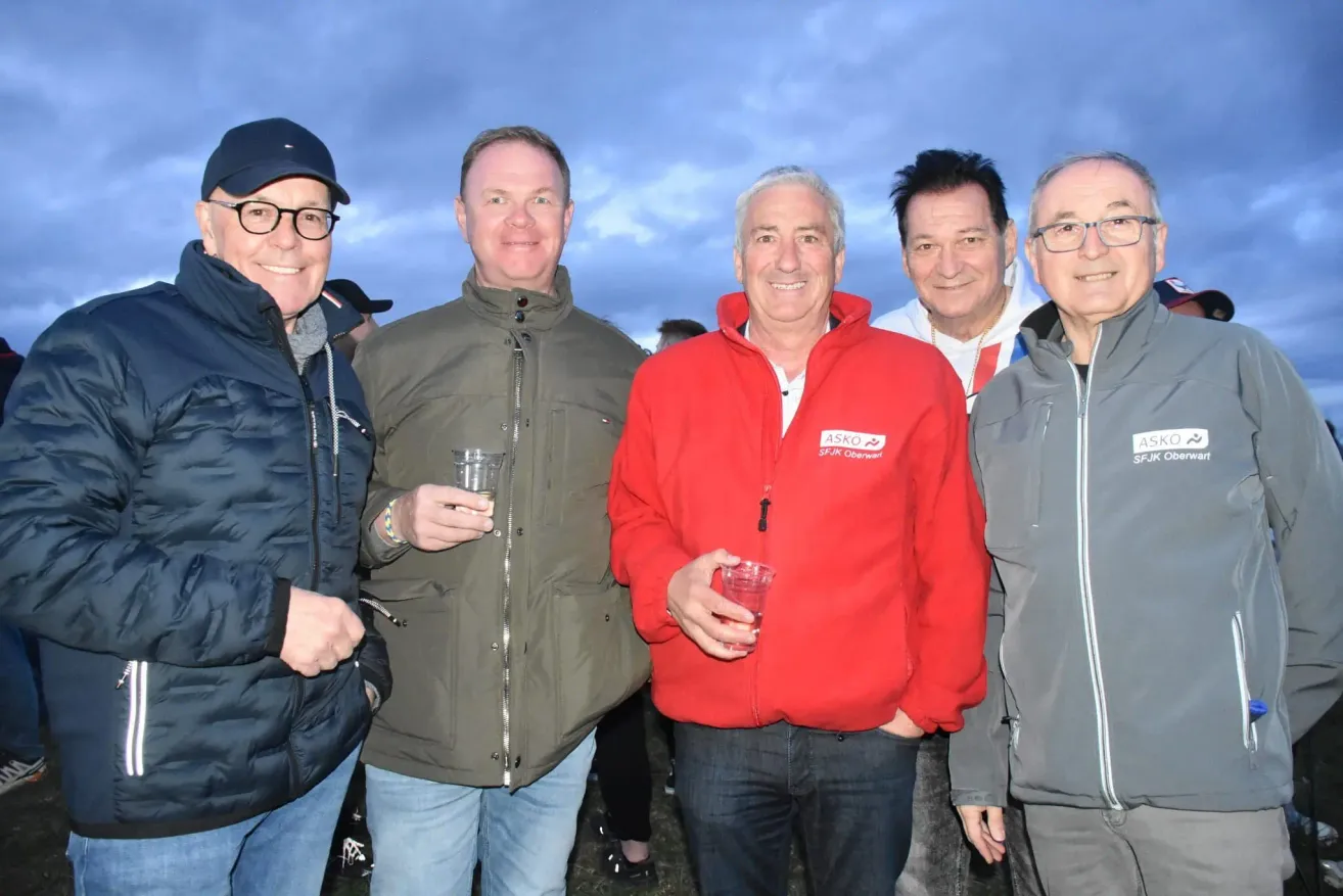 Five men stand close together, smiling. The man in the red jacket has a tag on his jacket that reads 'ASKO SFJK Oberwart'. The others are dressed in jackets and are holding drinks. They are all looking at the camera.