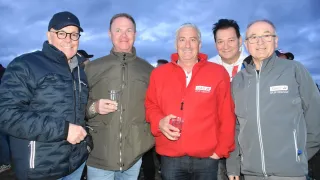 Fünf Männer stehen eng beieinander und lächeln. Der Mann im roten Jackett hat ein Etikett auf seiner Jacke, das 'ASKO SFJK Oberwart' besagt. Die anderen sind in Jacken gekleidet und halten Getränke. Sie alle schauen in die Kamera.