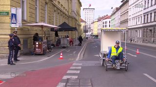 A man in a reflective vest sits on a tricycle near a bike cafe. Buildings and traffic cones line the street.